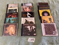 Collection of 14 music CDs from various artists arranged in rows on a fabric surface, showing front covers mostly intact. Includes Cowboy Junkies, Van Morrison, and multiple other albums. A paper labeled '25' is placed in front.