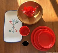 Overview of red plastic plates stacked, red plastic utensils in wooden bowl, and rectangular white tray with arrow graphic.