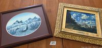 Two framed prints placed side by side on a wooden surface. Left: brown wooden frame with oval print of a woman by a pond and houses, attributed to Catherine Karns Munn. Right: rectangular ornate gold frame containing Claude Monet 'The Market at Argenteuil' print with visible label.
