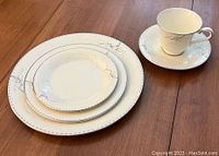 Stack of dinner plate, salad plate, bread and butter plate, and a cup with saucer in the background showing the Angela pattern with gray floral branches and beaded rim detail.