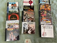 Photo shows a total of 17 CDs laid out in three groups on a green fabric surface. The CD titles include Dumb and Dumber, Angel, Pulp Fiction, Dawson's Creek, Billboard, Divas Las Vegas, Teenage Rock, 80s Rewind, and Sixties hits among others.