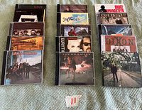 Top-down view of 16 CD cases arranged in a grid on a green cloth, showing album cover art from various well-known artists including Bruce Springsteen, Greg Allman, Genesis, and others.