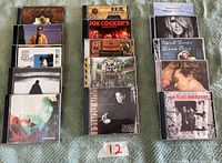 Photo of 15 music CDs by various artists arranged in three vertical stacks on a fabric surface, includes visible album covers showing artists names and titles.