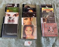 Photo showing 18 music CDs arranged in three columns on a blanket. Albums include INXS Greatest Hits, Eric Clapton, Johnny Cash The Essential, Diana Ross, John Arlen among others.