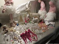 Photo showing three plush Christmas gnomes standing behind an arrangement of assorted Christmas decorations including a glittery gold reindeer, a white elephant figurine, oval light, and many ornaments.