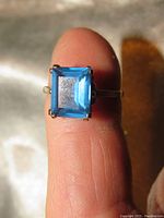 Close-up photo showing the vibrant square emerald cut blue sapphire set in 14K yellow gold finish on the ring, displayed on a finger to indicate size.