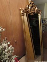 Full view of the tall ornate mirror showing gold-tone frame and reflective glass surface leaned against a wooden door.