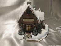Front view of Thomas Kinkade Hawthorne Village St Nicholas Christmas Shop model showing snow-covered roof, colorful Christmas lights, stone entrance, and two evergreen trees.