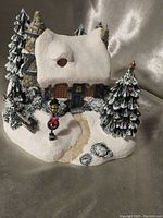 Front view of the Thomas Kinkade Stonehearth Hutch village house showing snow-covered roof, front door, window, path, pine tree, and wreath on lamp post.