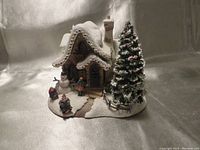 Angled front view showing cottage with snowman, girl figure, Santa, and large decorated evergreen tree on right side. Glittery snow cover and multi-colored light decorations on roof edges visible.