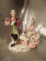 Front view of porcelain musical couple figurine showing man playing flute and woman at piano with lace dress under spotlight.
