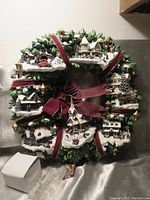 Front view of the ceramic Christmas Village Wreath showing detailed miniature snowy houses, lamp posts, and red velvet ribbons forming a decorative bow.