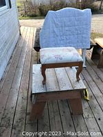 Decorative wooden footstool sitting on a wooden platform outdoors. The footstool has curved queen anne style legs and an upholstered seat with a floral patterned fabric.