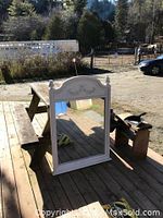 Front view of large decorative mirror showing carved floral detail on arched wooden frame painted in light color. Positioned on wood deck outdoors.