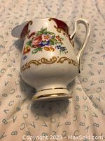 Small vintage cream jug with floral and gold accent decoration, lying on patterned cloth.