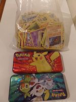 Clear large ziplock bag filled with many Pokemon trading cards and two metal tins, one with Pikachu on a red background and one with three Pokemon on blue/green background.