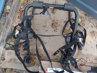 Top-down view of a black metal bike rack laid on cardboard with leaf debris around. Shows foam padding on bars and several attached black adjustable straps with buckles and hooks.