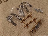 Photo of five black curved cabinet pulls individually packaged, three gold-tone bar style pulls also packaged, and screws, all on carpeted floor.