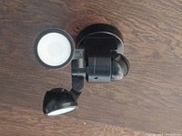 Black exterior light fixture with two LED lamps and a central motion sensor on a wood floor.