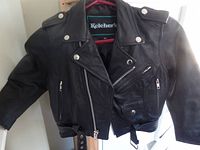 Front view of kids' black leather biker jacket on hanger showing asymmetrical zipper, three zippered pockets, belt and epaulets.