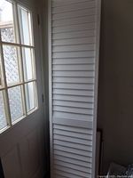 Full view of white plantation-style bifold door standing upright near window area, showing slatted design and panel frame.