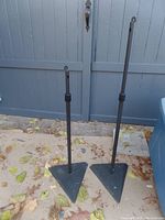 Two adjustable black metal speaker stands with triangular bases placed on the ground outside near a blue-gray fence.