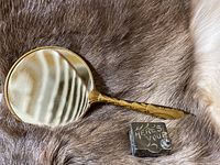 Gold-toned metal framed double-sided hand mirror with curved handle and magnified side shown along with the engraved silver trinket box on a fur surface.
