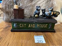 Front view of cast iron Cat and Mouse mechanical bank showing cats and mouse figures on a green base with 'CAT AND MOUSE' painted in gold.