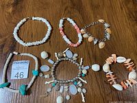 Overview of six bracelets on wooden surface showing mix of white shells, turquoise stones, and metal charms.