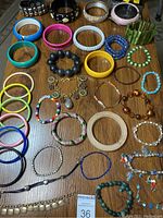 Photo shows 35 assorted bracelets laid out on a wooden surface, featuring a variety of materials such as plastic bangles, wooden bangles, beaded bracelets, and metal studded bracelets.