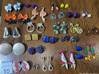 Photo showing 25 pairs of assorted earrings arranged on wooden surface, featuring various styles including studs, dangles, and clips in multiple colors and materials.