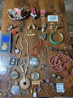 Full collection of costume jewelry, small decorative items and figurines arranged on wood surface, showing variety including keychains, medals, necklaces, bracelets, pendants, pins, and more.