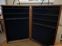 Photo showing the open jewelry showcase with two vertical sections, each having three horizontal shelves covered in black velvet and metal hooks beneath for hanging jewelry. Braided carry handle on top edge is visible.