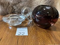 Two Kosta Boda glass candle holders displayed on wood surface, one clear with ring handle, one dark amethyst swirl glass sphere.