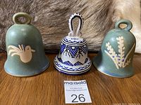 Front view of three ceramic bells on wooden surface with animal fur background; two green Norwegian bell cups with cream motifs and one white and blue Italian ceramic bell.