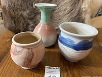 Three pottery pieces on wooden surface with fur background; small marbled pot, gradient glaze vase, and wide bowl-shaped pot with blue brushstrokes.
