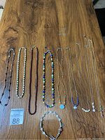 Full view of nine pieces of jewelry laid on a wood surface showing a mix of necklaces and one bracelet featuring various beads and silver chains.