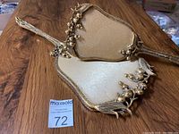 Photo of the brush and handheld mirror laid on wooden surface showing their decorative gold-tone floral and leaf design and beige backing.