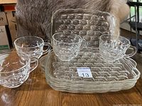 Four rectangular clear glass plates with bubble textures and cup grooves, accompanied by four matching bubble-patterned clear glass cups with handles, displayed on wooden surface.