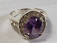 Close-up angled view of the oval amethyst ring showing purple center stone with surrounding clear stones and silver-tone band.
