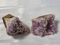 Two amethyst mineral specimens with purple crystals on rough rock bases, displayed on a light surface.