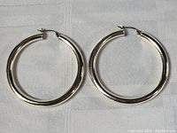 Pair of large sterling silver hoop earrings laid flat on white woven fabric background, showing full round shape and polished silver surface.