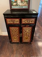 Front view of black Asian style cabinet showing two drawers and double doors with gold and red carvings, visible paint wear and finish damage.