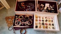 Overview of assorted costume jewelry displayed in boxes and trays showing necklaces, bracelets, rings, earrings.