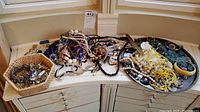 Wide view of costume jewelry lot including necklaces, earrings, bracelets arranged in basket and metal pan
