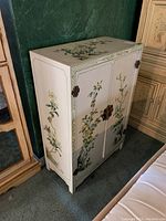 Full exterior view of white cabinet with floral designs on sides, doors, and top. Shows overall shape and decorative details.