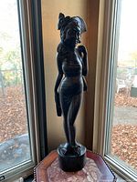 Full view of the 23 inch tall hand carved wooden female figurine standing on a base in front of a window.