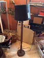 Floor lamp shown in a cluttered basement area, displaying full view of lamp with black faux suede shade and wood base.