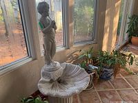 Overall view of the figural fountain showing the child figure standing atop a shell-shaped basin, placed indoors near windows with surrounding plants.
