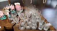 Overview of arranged glassware including stemmed glasses, etched glasses and tall plain glasses, plus pink flamingo figurines and a pink pitcher.